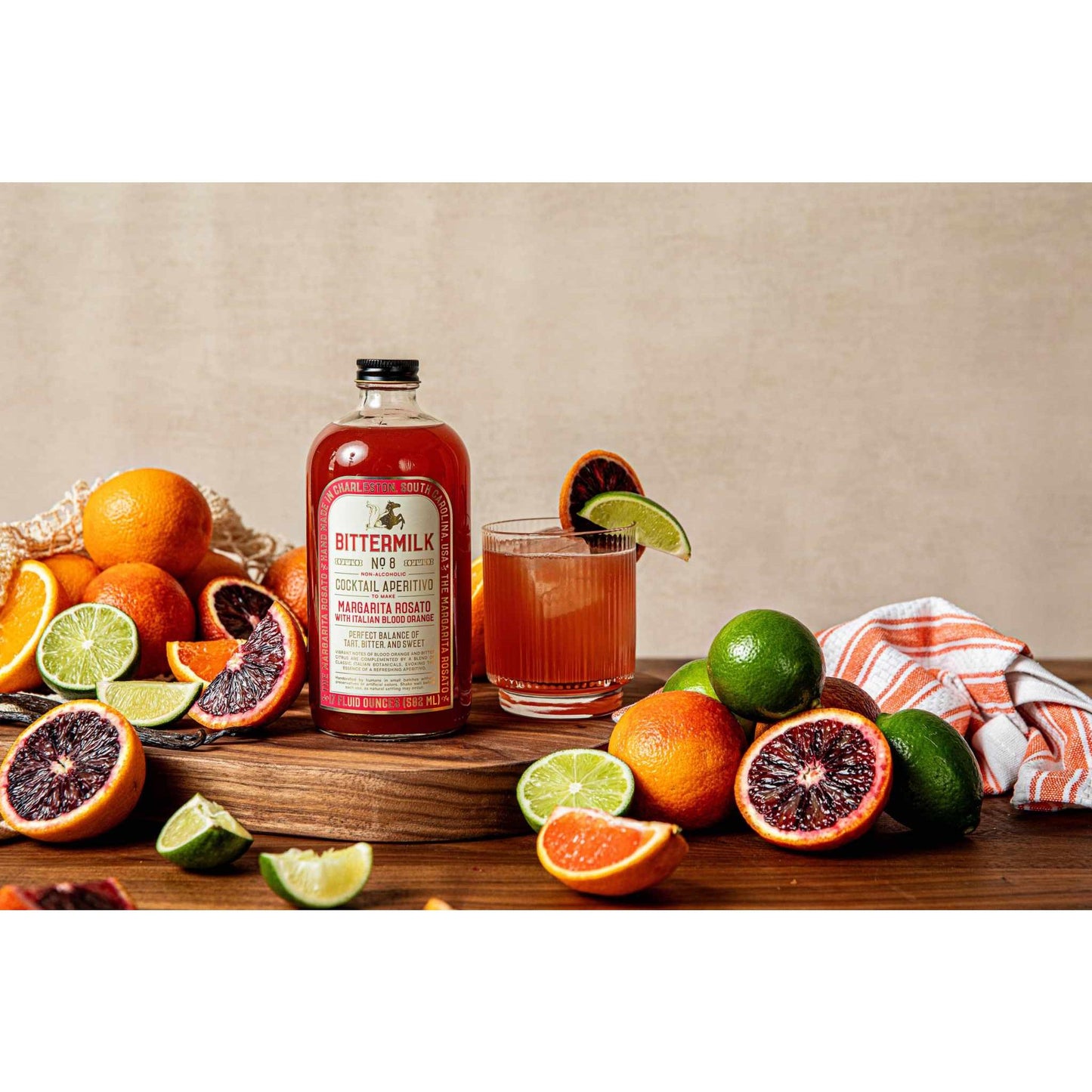 Bittermilk No.8 Margarita Rosato mixer bottle with fresh blood oranges, limes, and a glass of cocktail garnished with citrus slices on wooden surface.