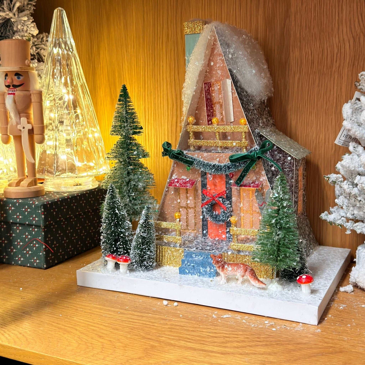 
                  
                    Handcrafted glittered A-Frame Cabin with pre-lit windows and bottle-brush trees by Cody Foster Co.
                  
                