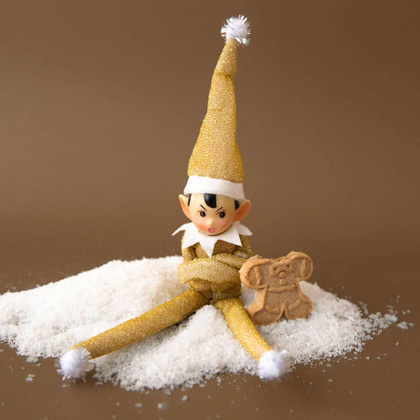 Gold glitter elf ornament with bendable limbs and pompom hat seated next to gingerbread cookie on fake snow