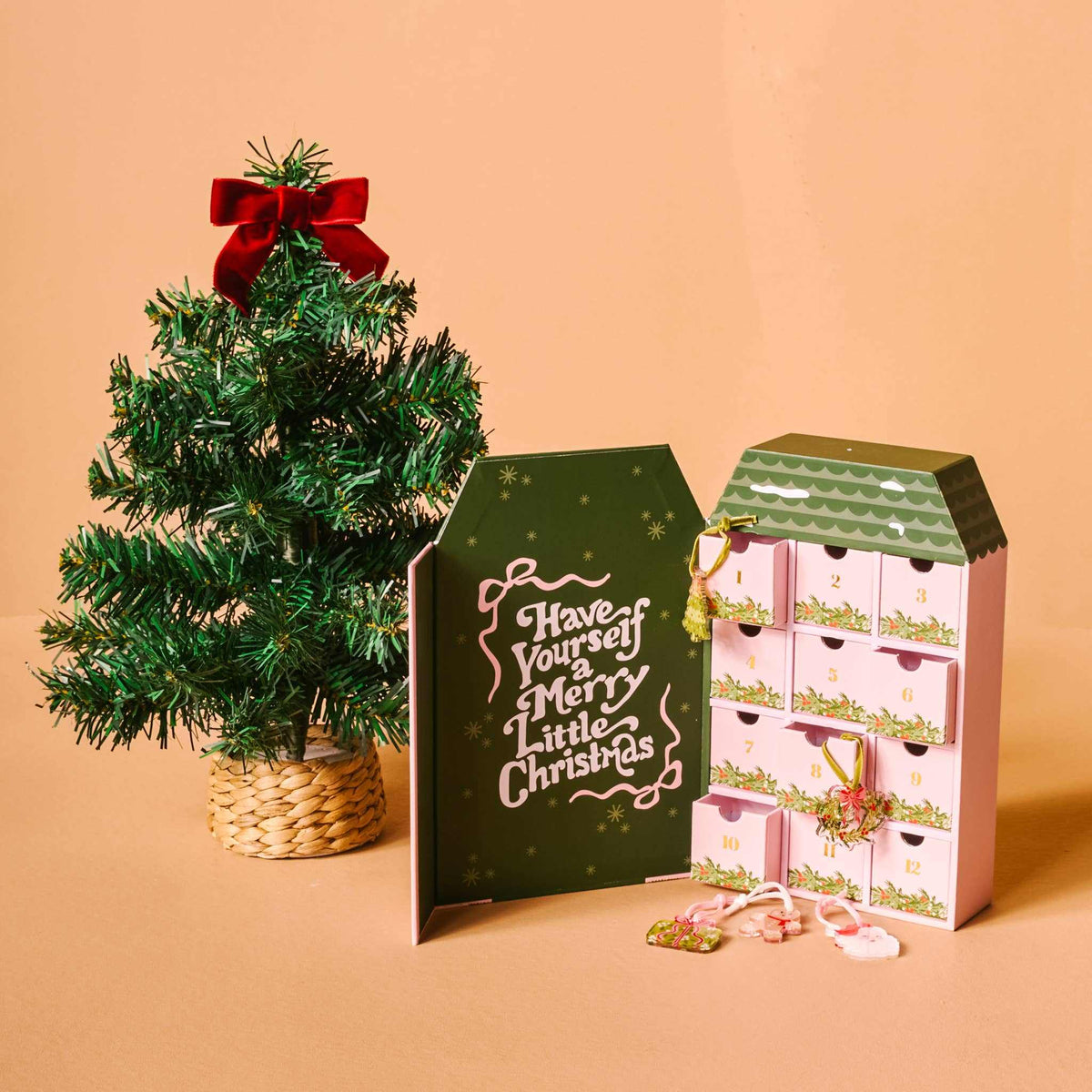 
                  
                    Mini artificial Christmas tree with red bow beside pink Advent Village House calendar with 12 miniature Christmas ornaments for holiday countdown
                  
                