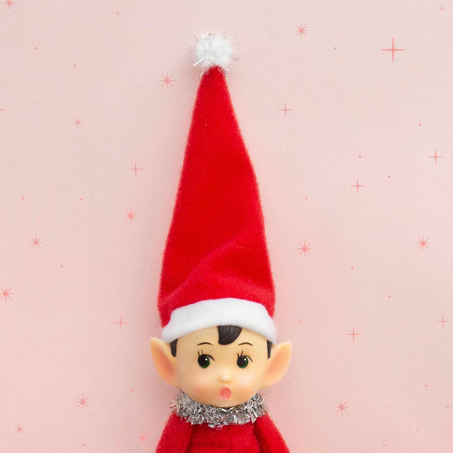 Festive Frankie elf ornament with red felt hat, tinsel collar, and bendable limbs on pink background