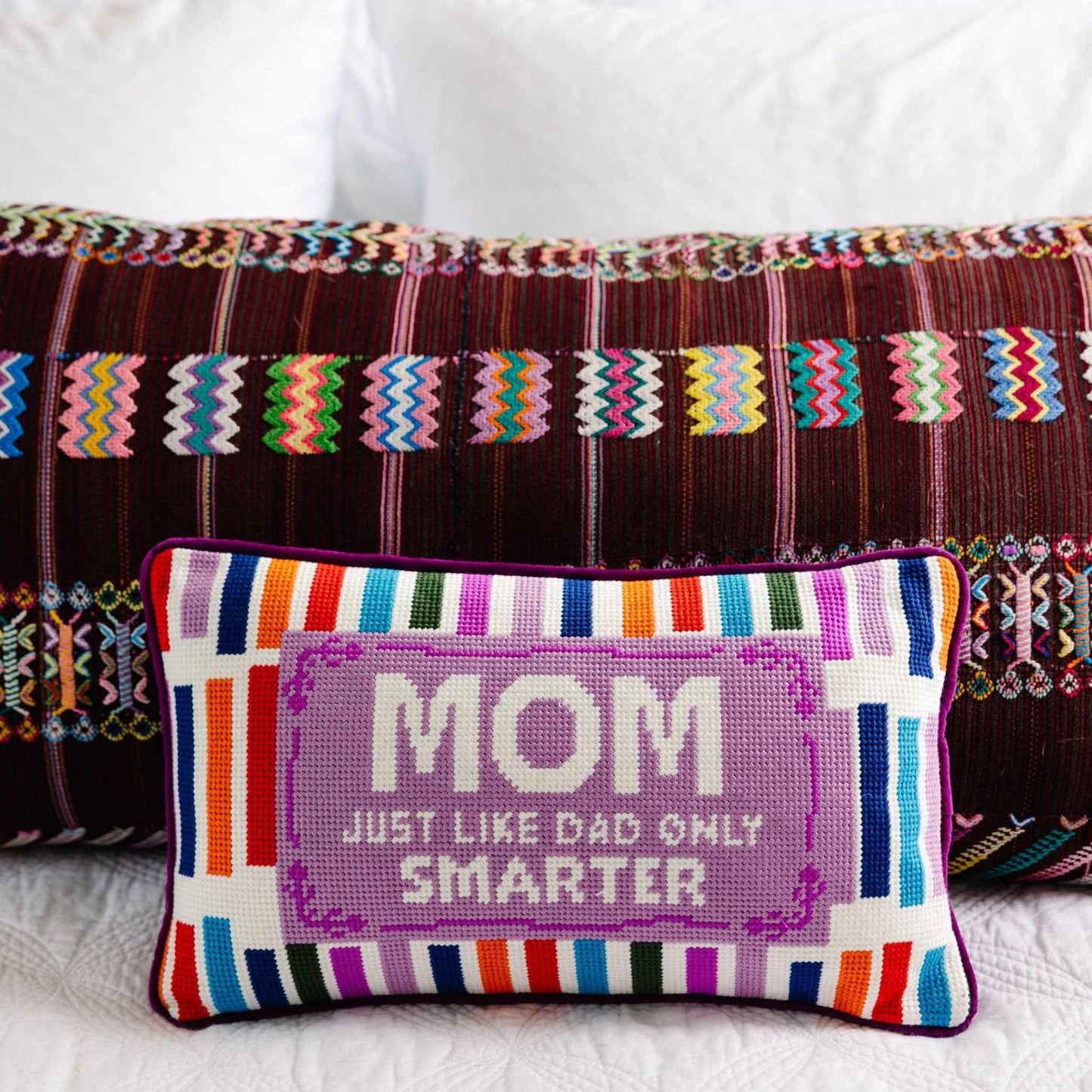 Mom Rules Needlepoint Pillow with colorful stripes and witty embroidered text on a bed with patterned pillows