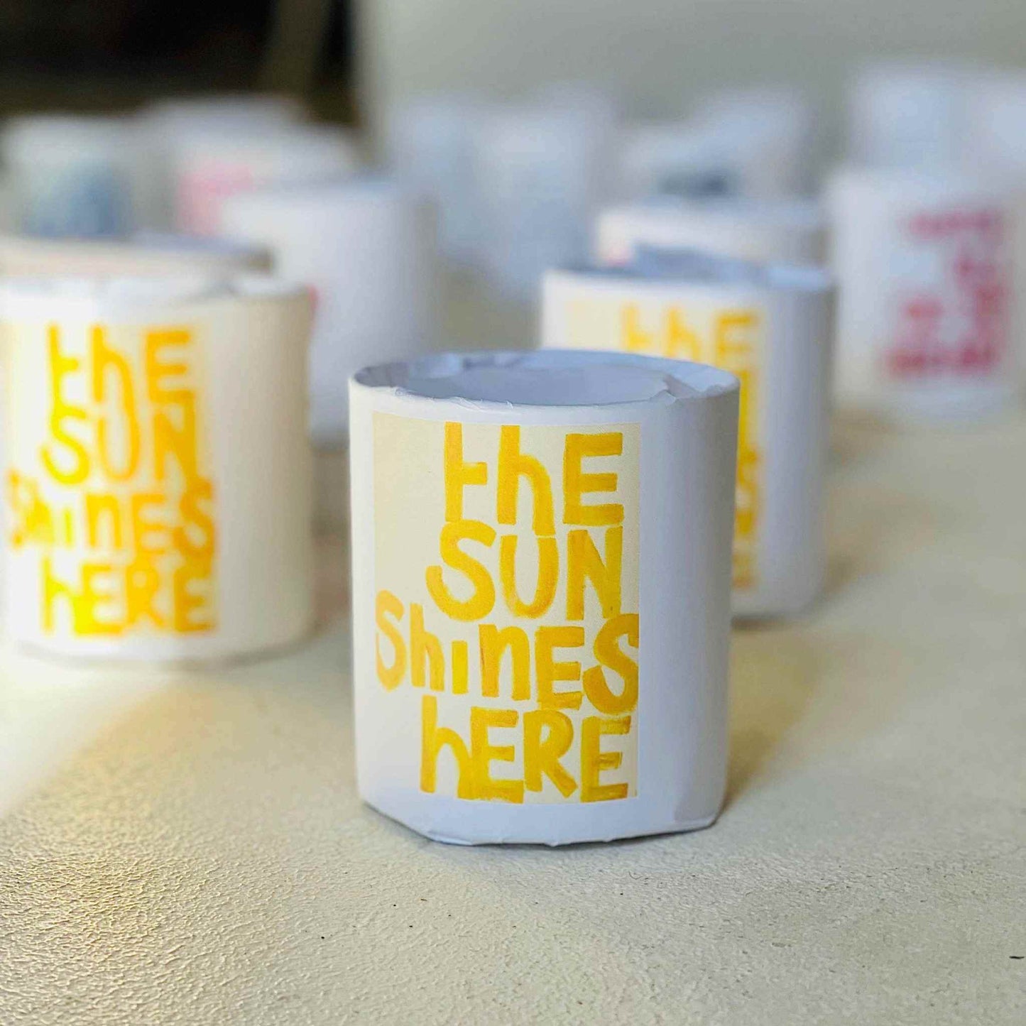 The Sun Shines Here soy candle with lemongrass, cedarwood, rose, and vanilla scent in white container with yellow text