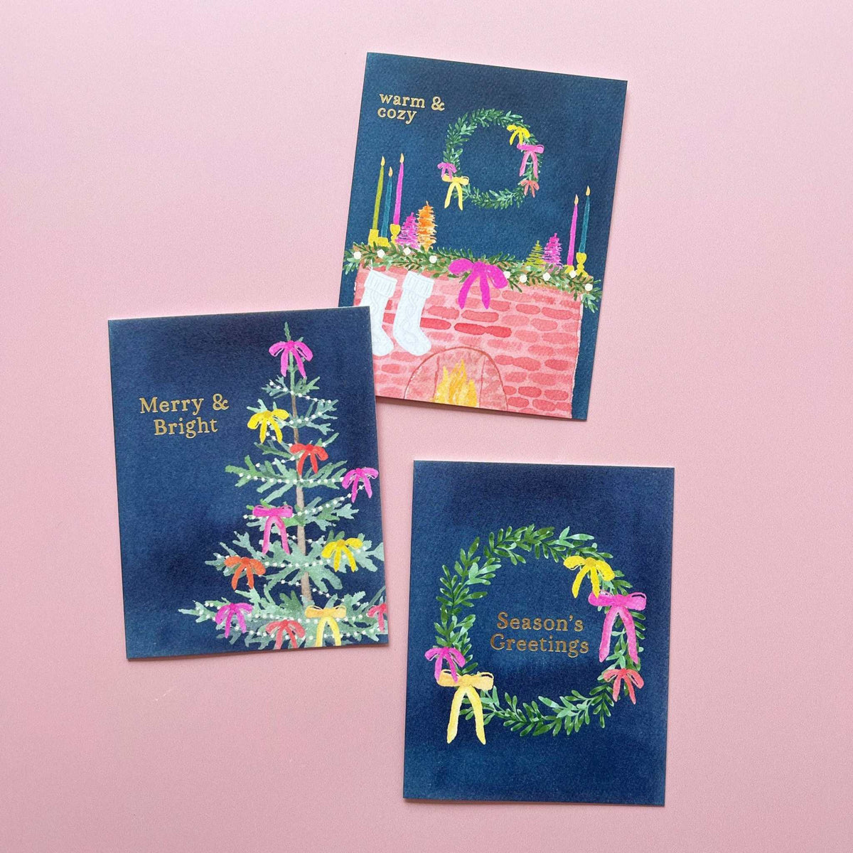 
                  
                    Holiday mantel greeting card with gold foil "Warm & Cozy" text, hand-painted mantel scene, festive wreath, bows, stockings on navy background
                  
                
