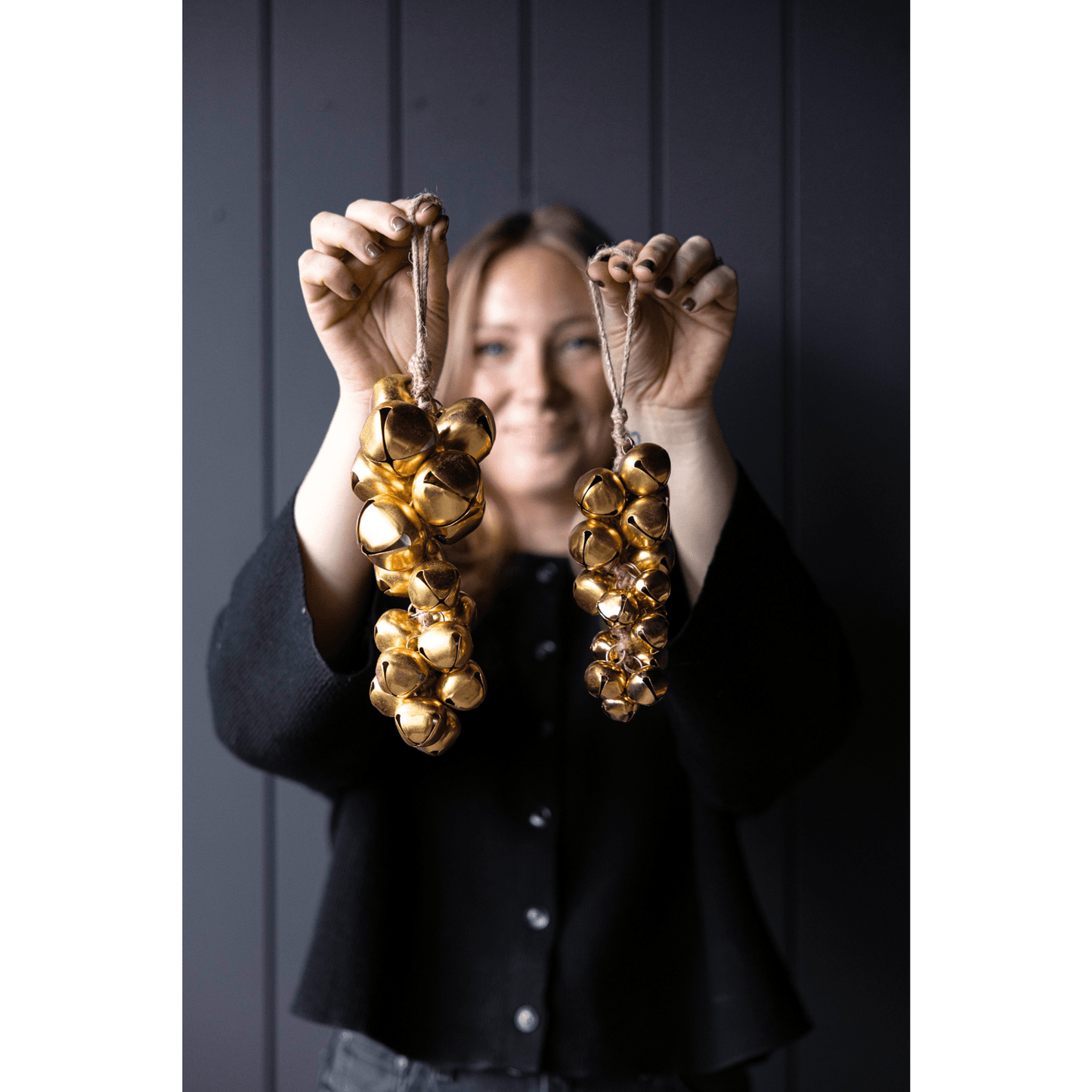 Person holding two gold jingle bell holiday hanging clusters against a dark background