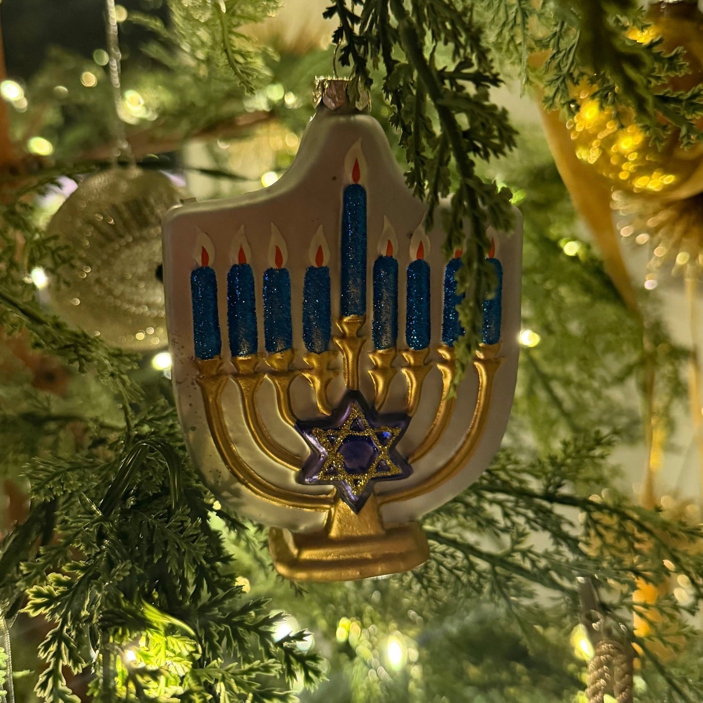 Glass menorah ornament with blue glitter candles and gold details hanging on a tree for Hanukkah decoration