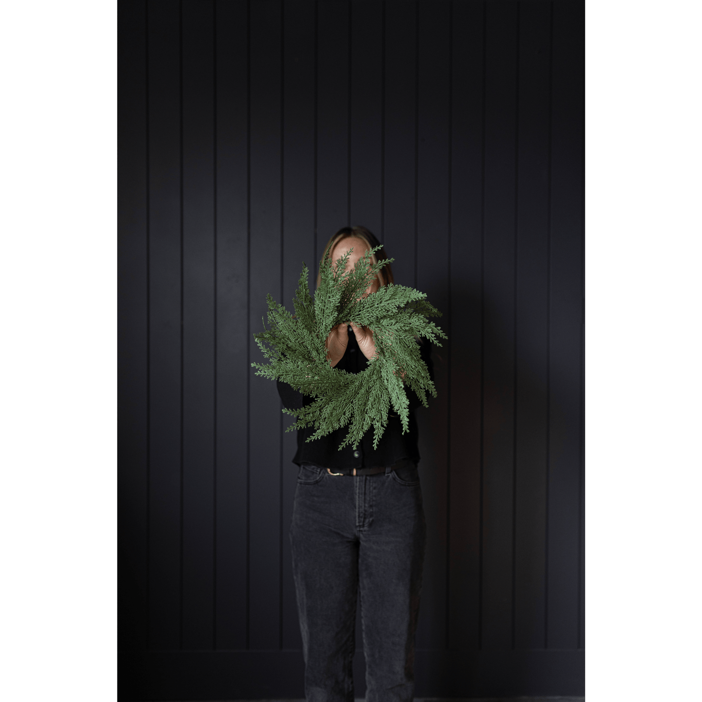 Woman holding 14-inch Cypress Leaf Full Real Touch Wreath with dense faux cypress foliage against black wall
