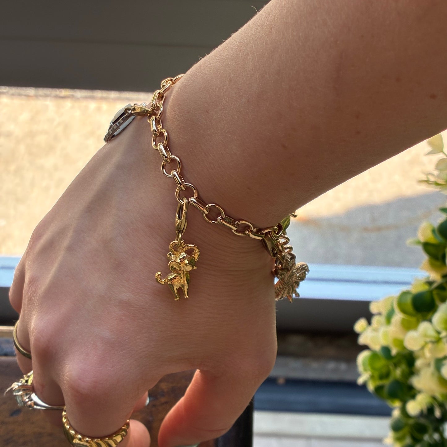 
                  
                    A gold chain bracelet with small dangling charms, worn on a person's wrist. The bracelet has a warm yellow-gold tone and is shown in a real-world setting with a hand and flowers visible.
                  
                