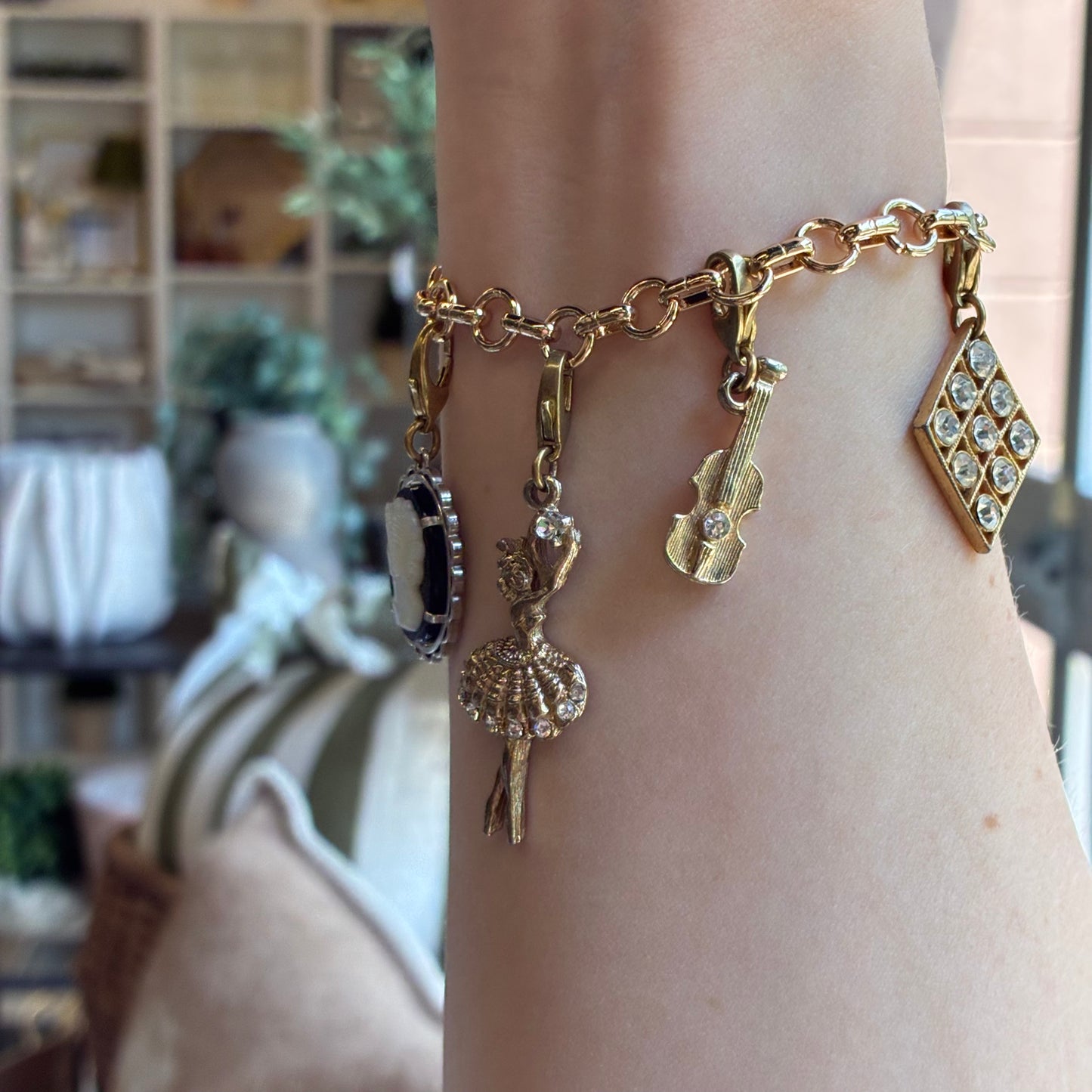 
                  
                    A gold charm bracelet with multiple dangling charms, including a ballerina, violin, and diamond-shaped crystal pendant, shown worn on a person's wrist.
                  
                