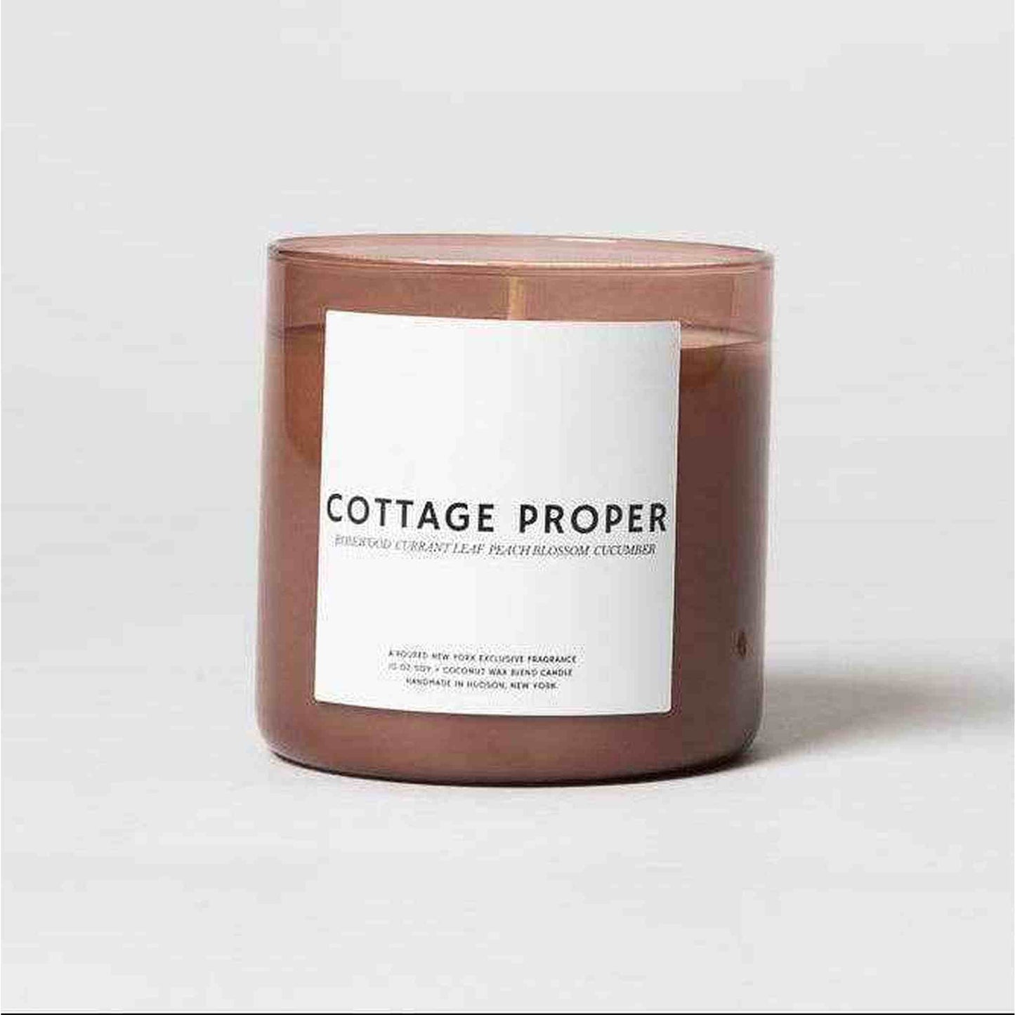 Cottage Proper Candle in a brown glass jar with a white label featuring rosewood, currant leaf, cucumber, and peach blossom fragrance.