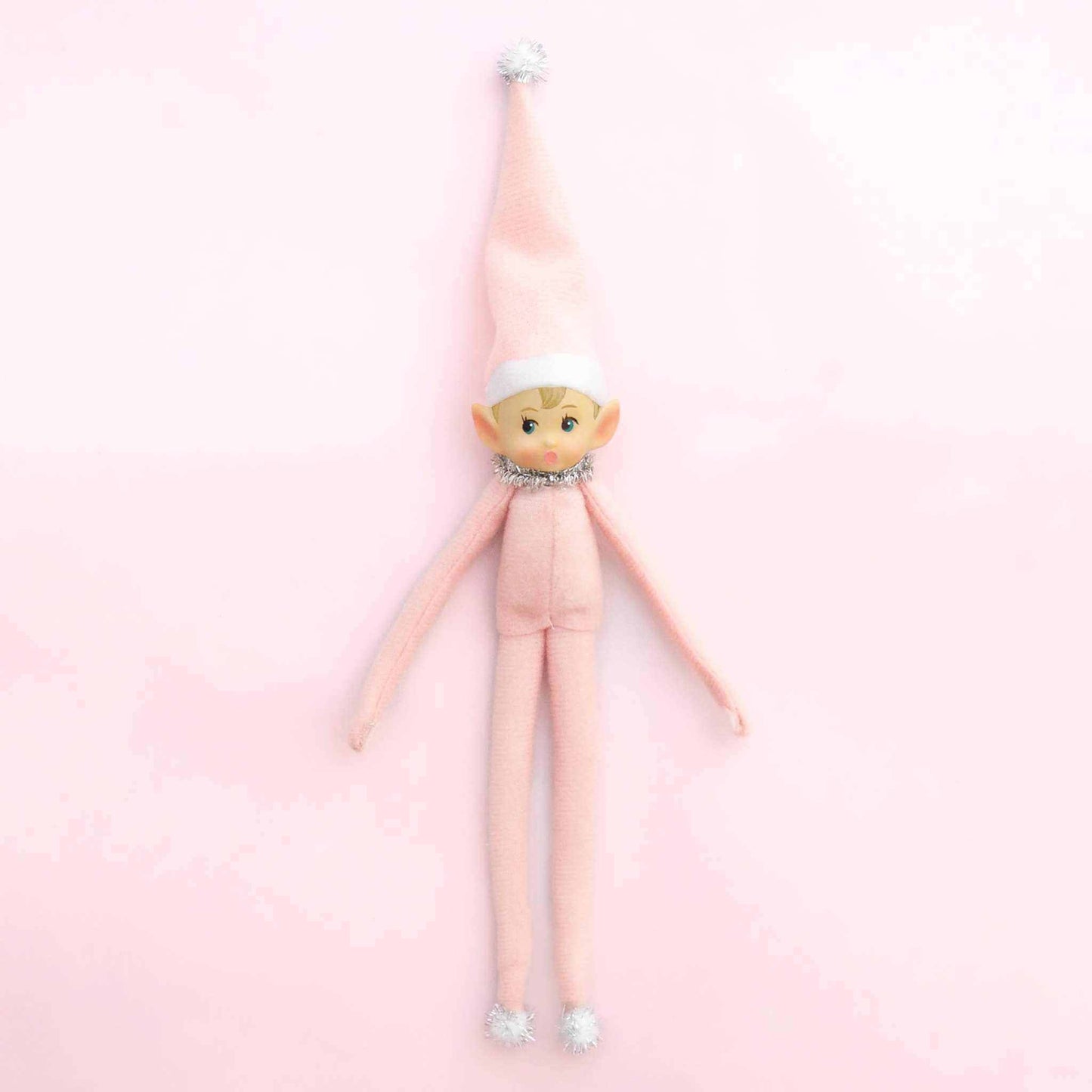 Elf ornament Little Lou with pink felt body, bendable limbs, and tinsel collar against a pink background.