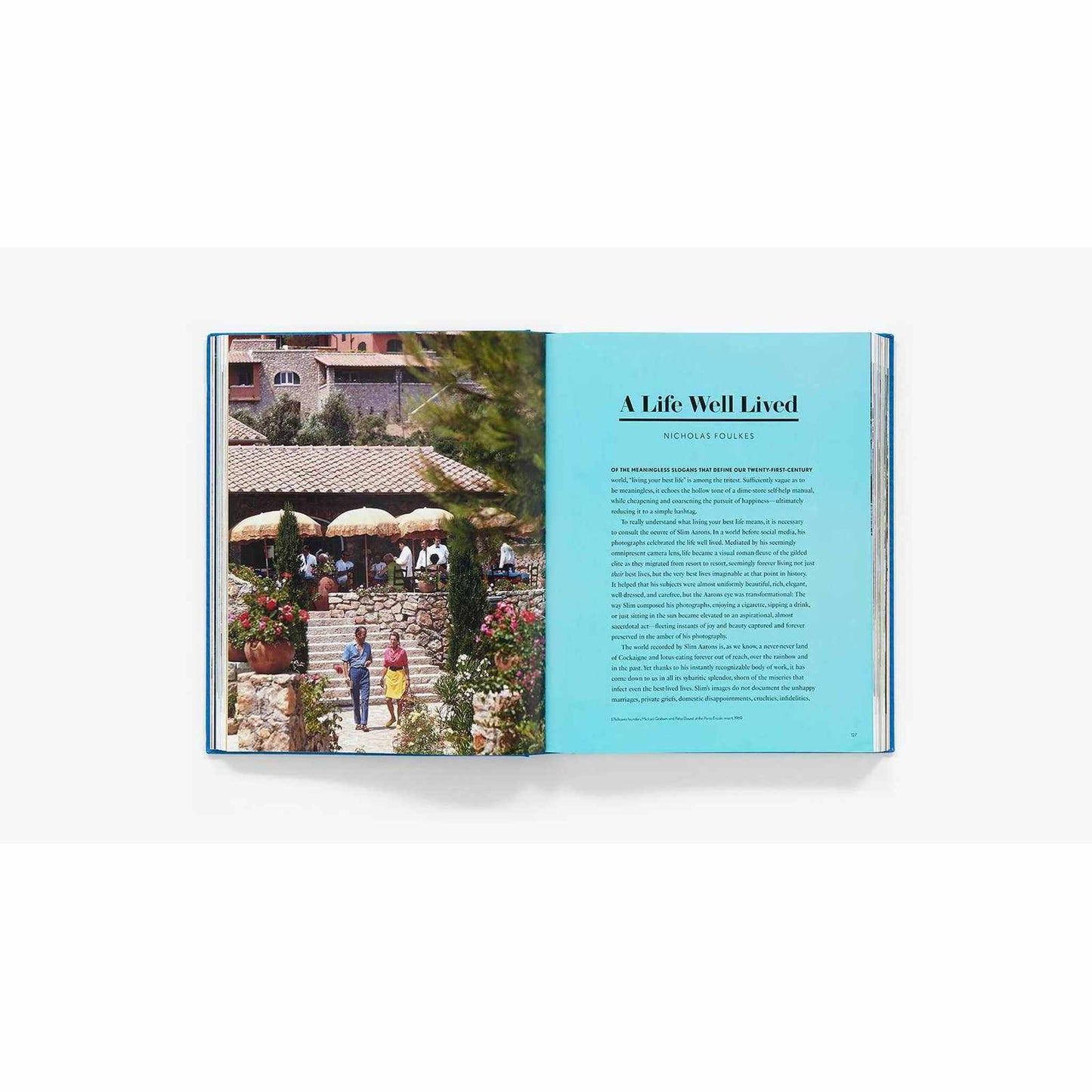 
                  
                    Open book showing a Slim Aarons photograph of a Mediterranean scene with people walking and outdoor umbrellas beside a biographical essay page titled "A Life Well Lived"
                  
                