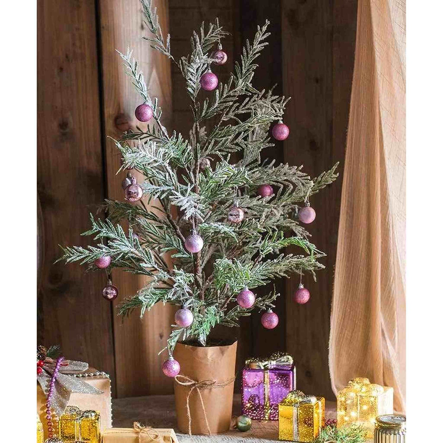 Snow flocked cedar fir tree with pink ornaments in kraft-paper wrapped base for indoor holiday decoration