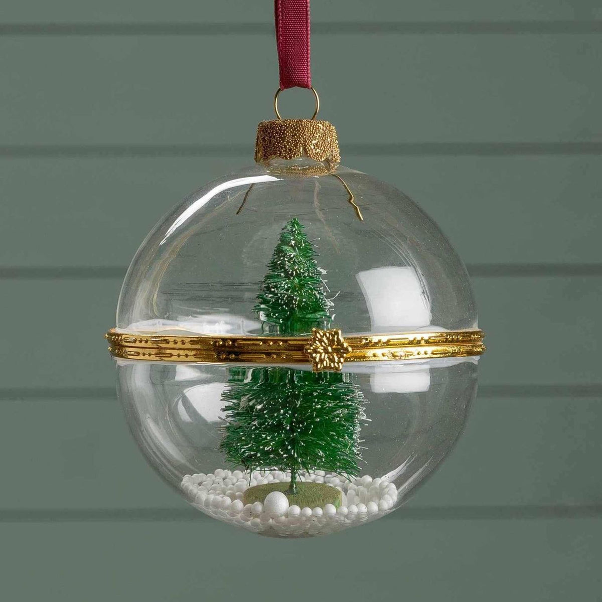 Clear glass ball ornament with miniature Christmas tree and faux snow inside, decorated with a gold band and red ribbon hanger.
