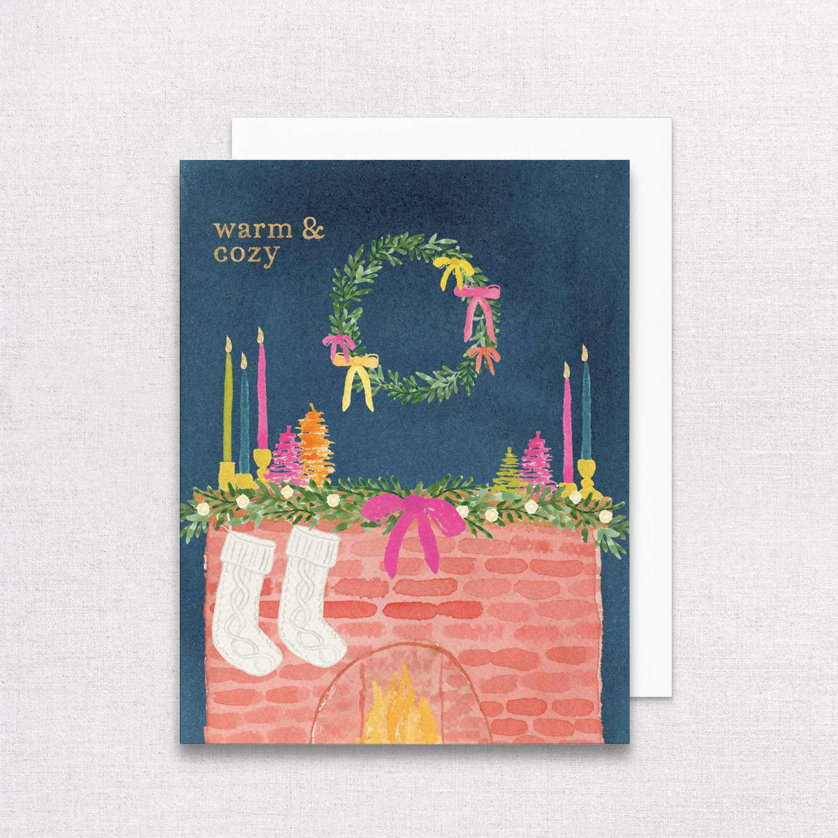 Holiday mantel greeting card with gold foil "Warm & Cozy" text, featuring a festive wreath, stockings, and candles on navy background