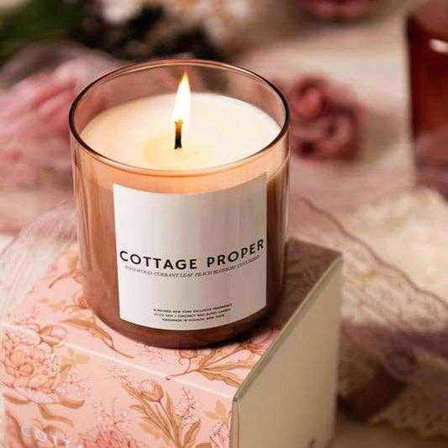 Cottage Proper Candle lit in a glass jar with floral patterned background and soft warm ambiance