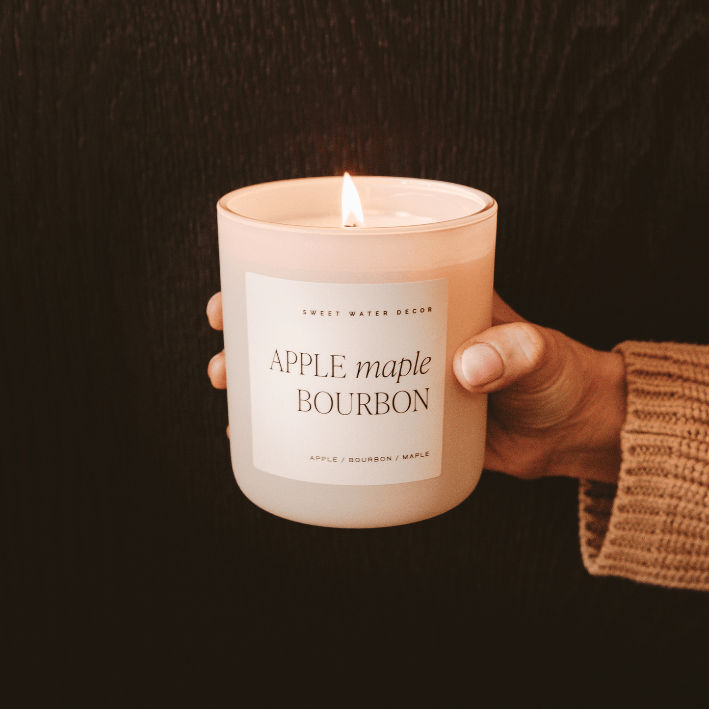 
                  
                    Hand holding lit Apple Maple Bourbon 15 oz soy candle with wood lid and fall-inspired label
                  
                