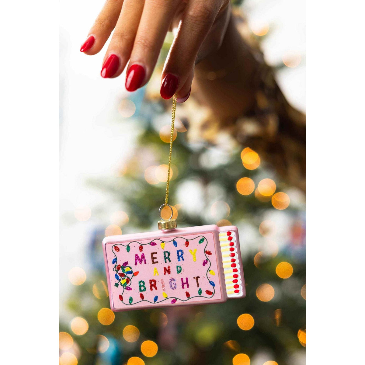 Hand holding Merry & Bright Matchbook Ornament with colorful lights and festive design against blurred Christmas tree background
