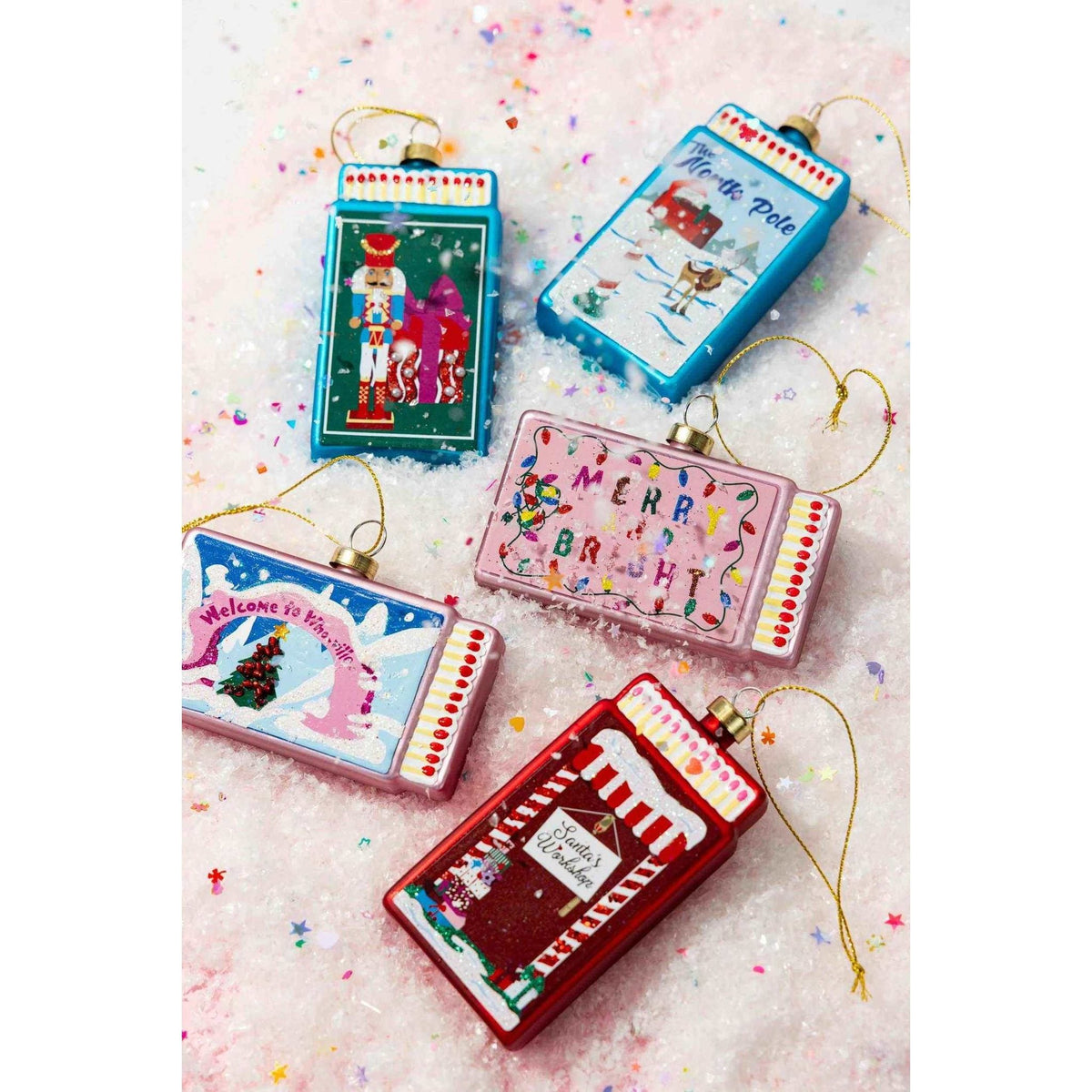 
                  
                    Handcrafted glass Santa's Workshop Matchbook Ornaments with festive holiday designs and gold hanging strings on a snowy background
                  
                