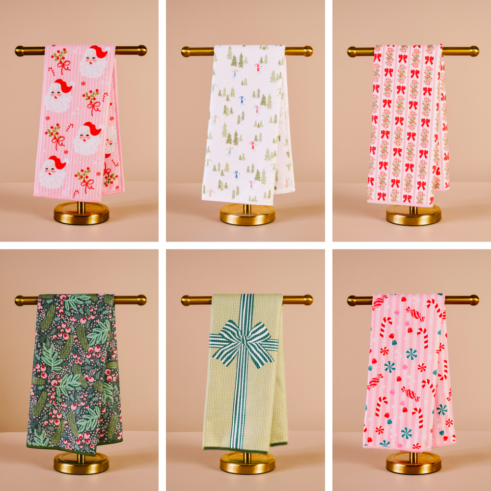 Holiday waffle tea towels with assorted festive patterns hanging on gold towel racks, featuring Santa, trees, candy canes, and holiday gift designs.
