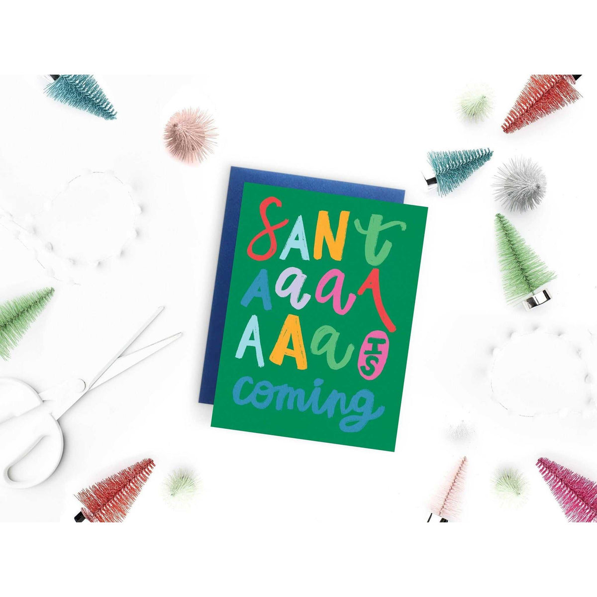 Santaaa Card colorful holiday greeting card with playful lettering on green background and blue envelope surrounded by festive miniature trees and craft scissors