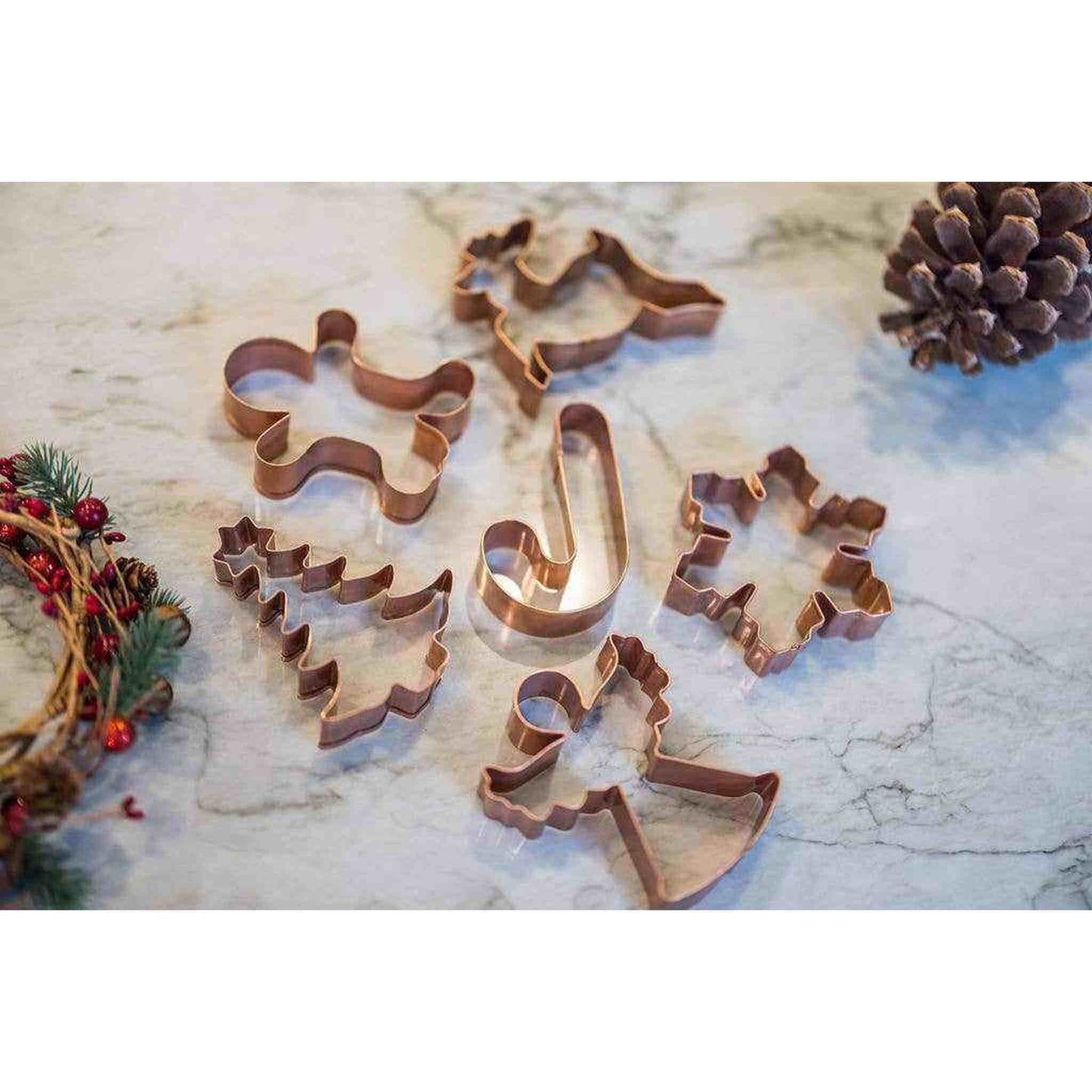 Set of six copper Christmas cookie cutters in tree, reindeer, candy cane, snowflake, angel, and gingerbread shapes on marble surface