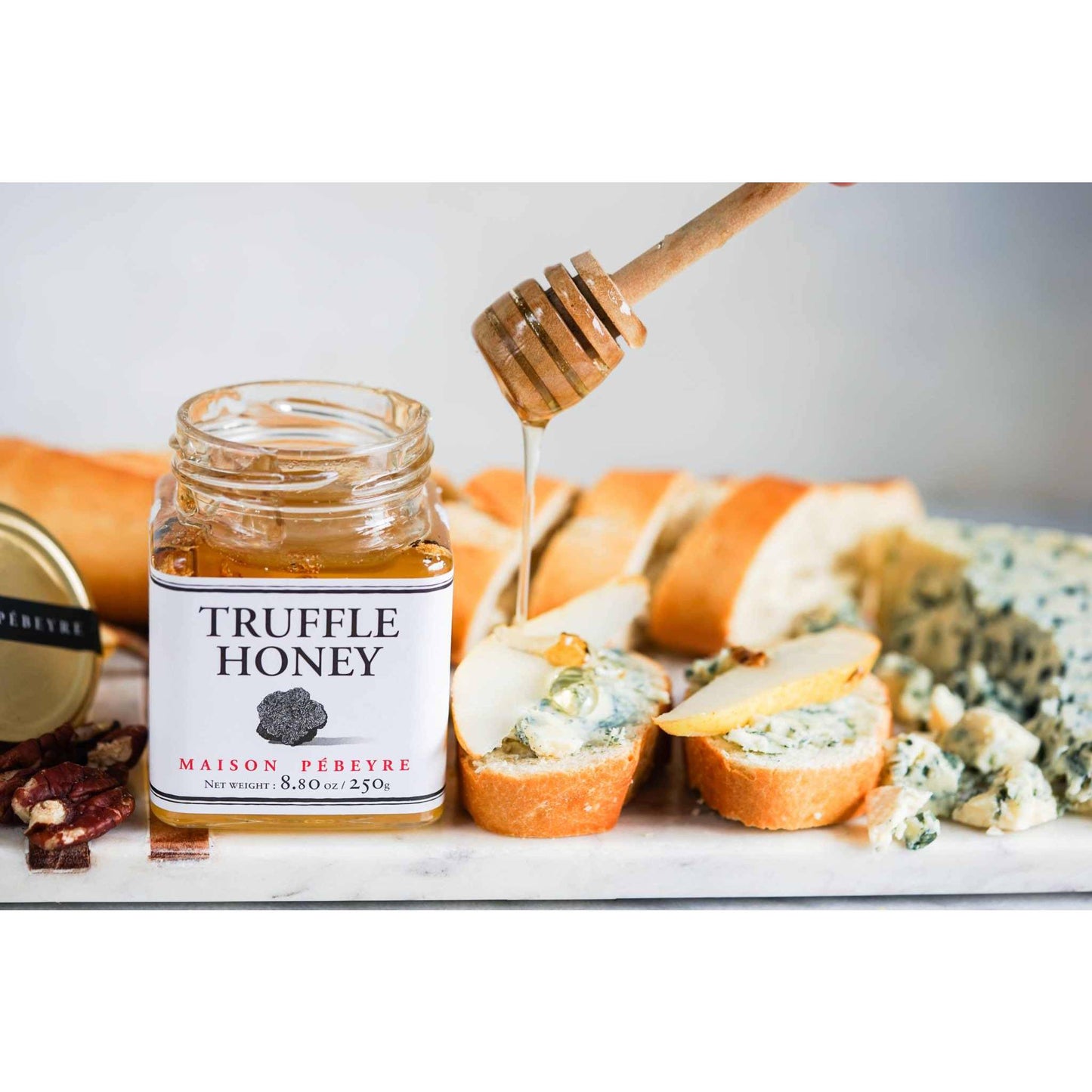 Jar of Pebeyre Truffle Honey with honey dipper drizzling over cheese and bread.