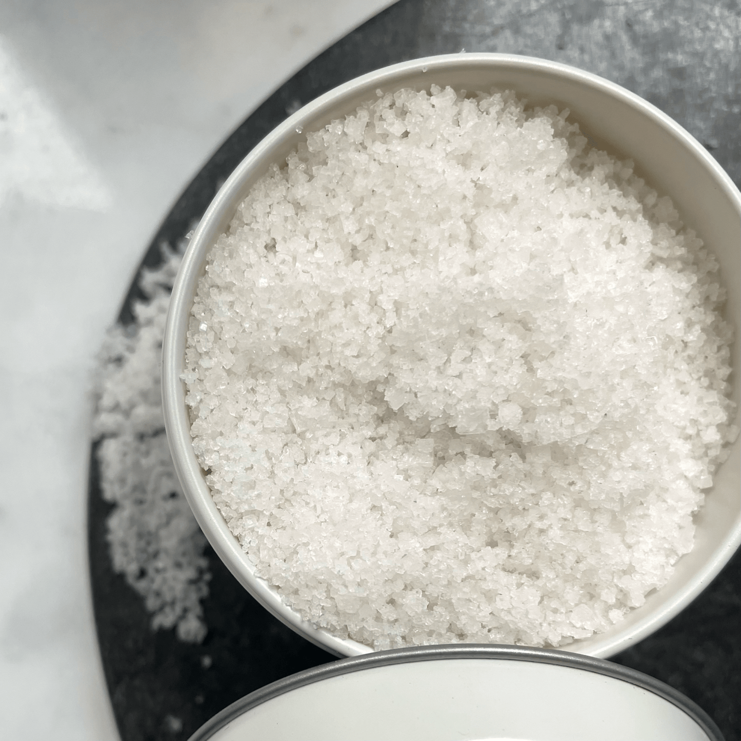 Close-up of Kampot Fleur de Sel sea salt crystals in a white container on a black surface