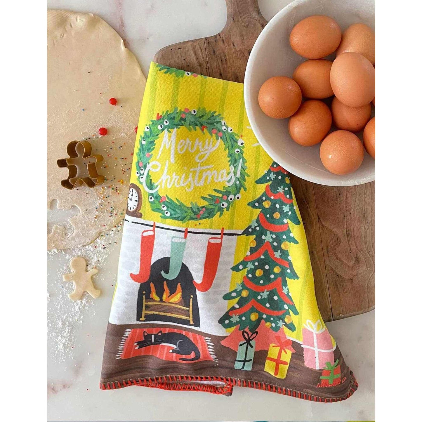 Festive Fireplace Tea Towel with colorful Christmas design, embroidered trim, and hanging loop on kitchen counter with baking items