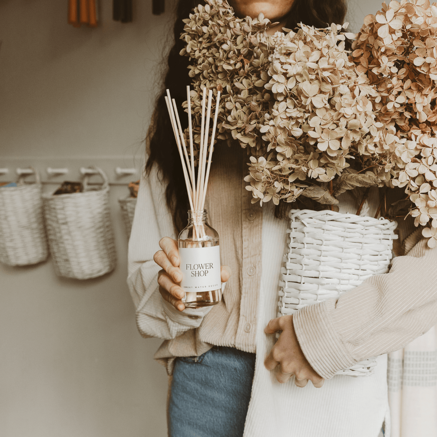 Flower Shop Reed Diffuser with natural reeds and fresh floral scent in clear glass bottle held by person with dried flowers and white basket.